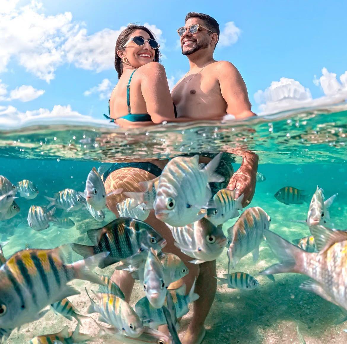 Casal sorrindo dentro das piscinas naturais de Porto de Galinhas, cercado por peixes coloridos e água cristalina